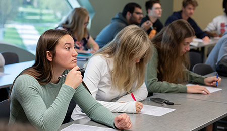 Students sit at desks in a classroom, working on assignments. In the foreground, one student in a green long-sleeve shirt holds a pencil near their face while looking ahead, with papers spread on the desk. Other students nearby are writing or working on laptops, and additional students sit in rows behind them.